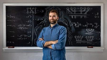 A graduate student, Andrew Adair, posing for a portrait with his arms crossed in a classroom. Behind him is a black chalkboard full of math equations written with white chalk.